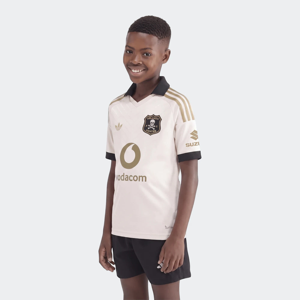 Young boy models the Orlando Pirates 26/27 Away Jersey for kids, featuring an off-white and gold design with black shorts.