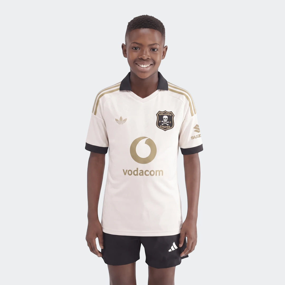 Young boy models an Orlando Pirates 26/27 Away Jersey in off-white, black, and gold, paired with black shorts.