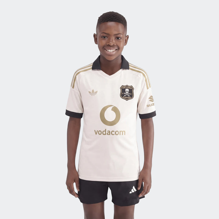 Young boy models an Orlando Pirates 26/27 Away Jersey in off-white, black, and gold, paired with black shorts.