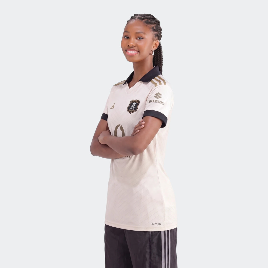 Young woman wearing an Orlando Pirates 26/27 Away Jersey in light cream with black and gold details, arms crossed.