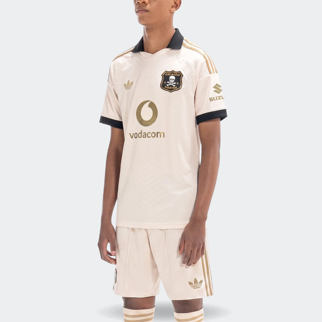 Model wearing Orlando Pirates 26/27 Away football kit, featuring a beige jersey, matching shorts, and socks with black and