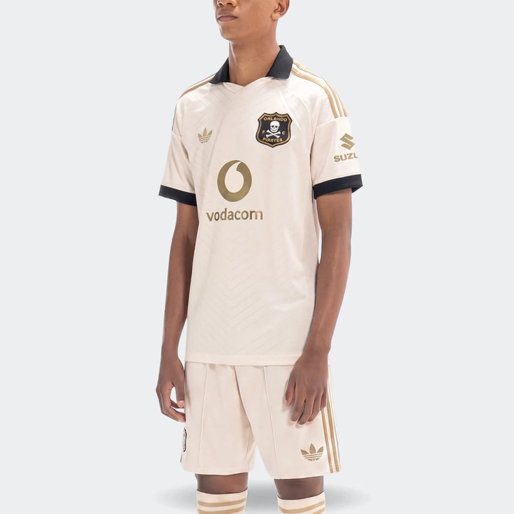 Model wearing Orlando Pirates 26/27 Away football kit, featuring a beige jersey, matching shorts, and socks with black and