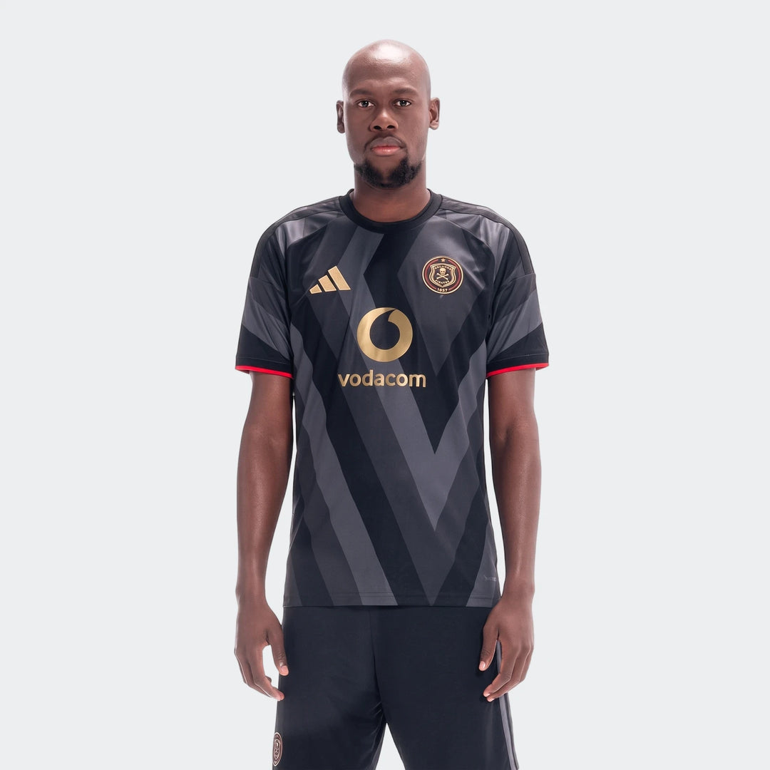 A bald man wearing the black and dark grey chevron Orlando Pirates 26/27 Home Jersey with gold Adidas and Vodacom logos.