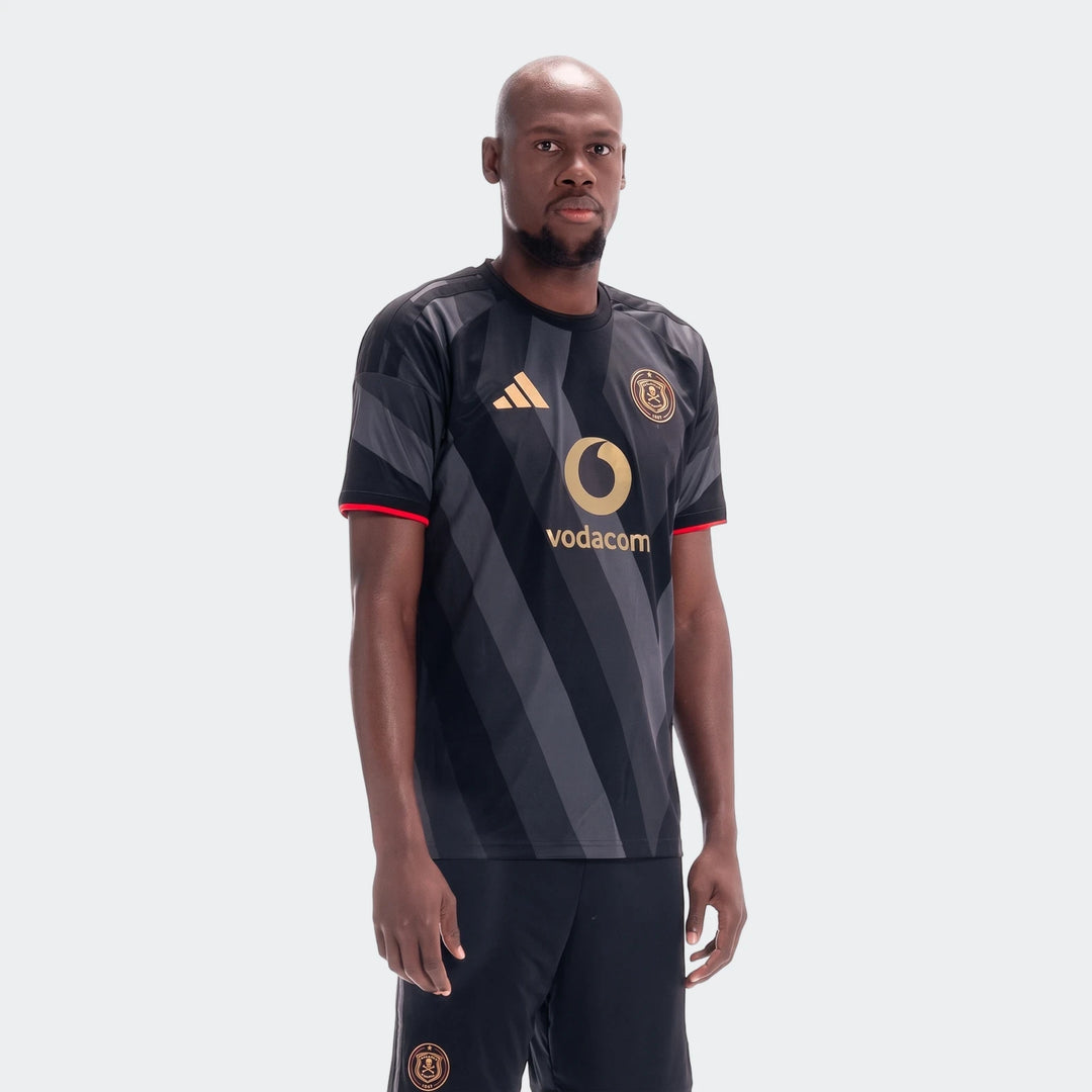 A male model wears the Orlando Pirates 26/27 Home Jersey, featuring black and grey stripes with gold logos and red trim.
