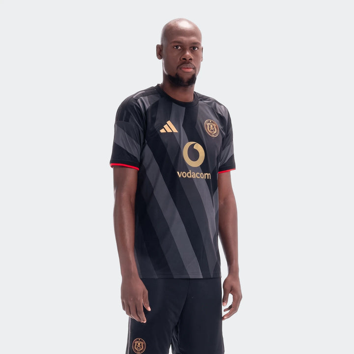 A male model wears the Orlando Pirates 26/27 Home Jersey, featuring black and grey stripes with gold logos and red trim.