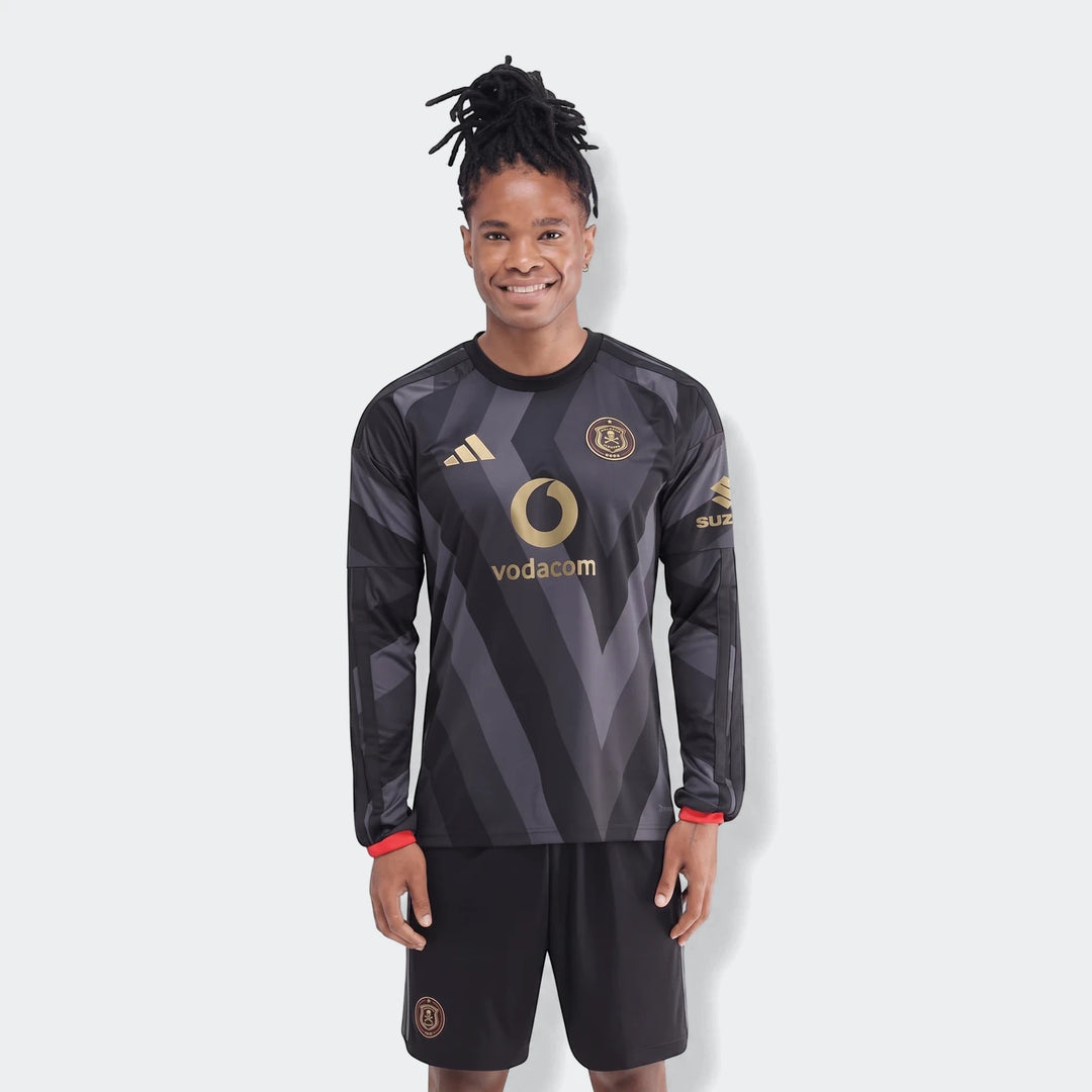 A man wearing the Orlando Pirates 26/27 Home Jersey, a black long-sleeved football shirt with gold logos, standing on a