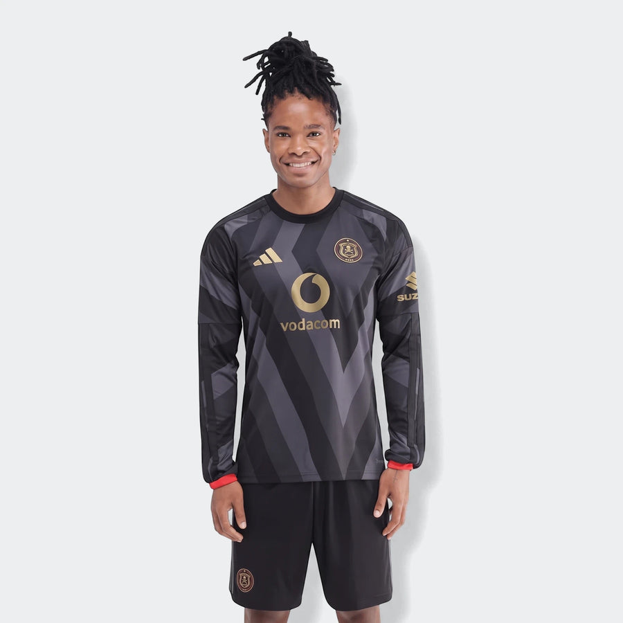 A man wearing the Orlando Pirates 26/27 Home Jersey, a black long-sleeved football shirt with gold logos, standing on a