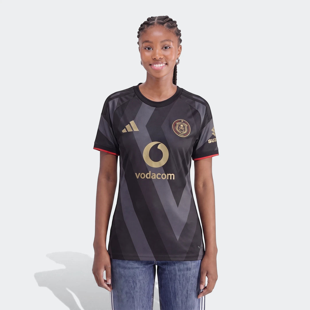 A smiling woman models the Orlando Pirates 26/27 Home Jersey, featuring black and grey stripes and gold sponsor logos.