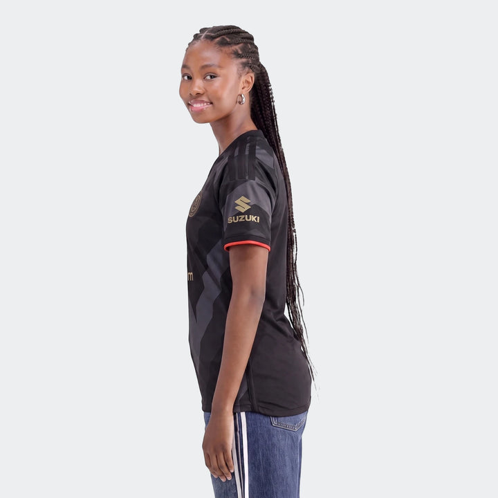 Smiling woman wears a black Orlando Pirates 26/27 Home Jersey with gold Suzuki logo, red trim, and blue jeans.