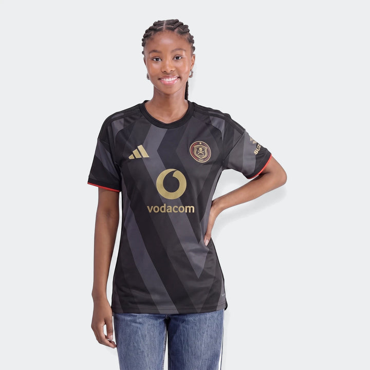 Young woman in a black and grey Orlando Pirates 26/27 Home Jersey with gold logos and red trim, smiling against a white