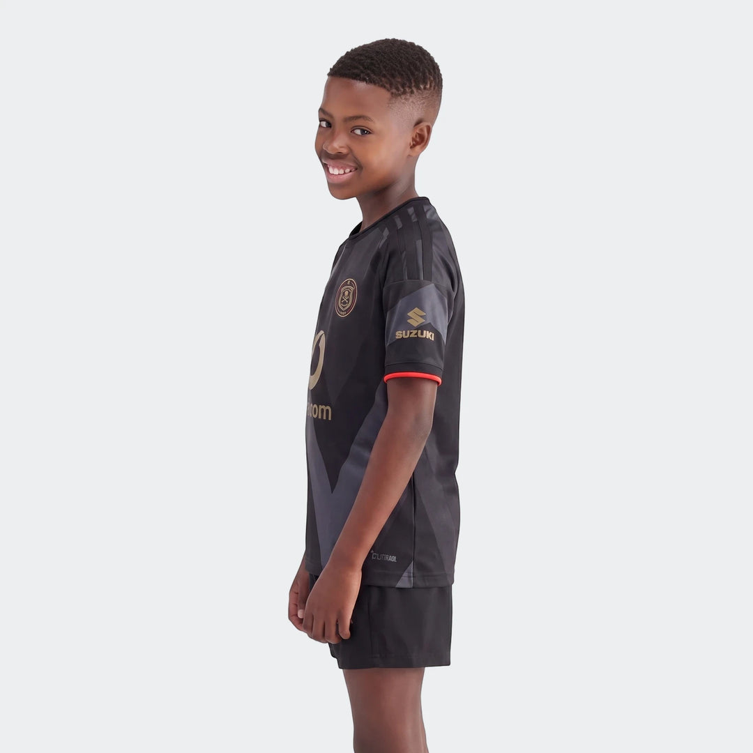 A young boy smiles while wearing the black Orlando Pirates 26/27 Home Jersey with gold logos and black shorts, in a side