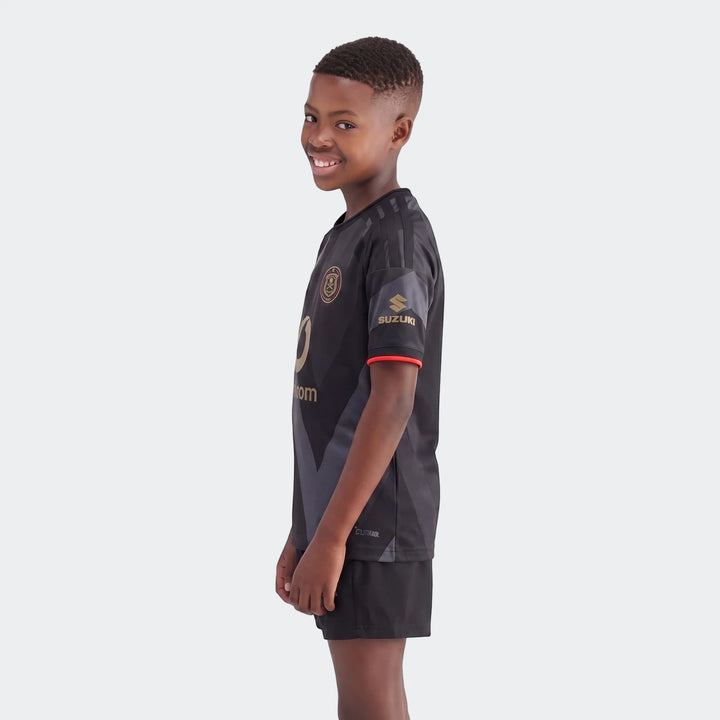 A young boy smiles while wearing the black Orlando Pirates 26/27 Home Jersey with gold logos and black shorts, in a side