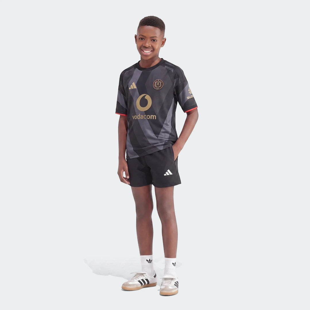 Young boy wearing a black and gold Orlando Pirates 26/27 Home Jersey, black shorts, and white sneakers, standing full body.