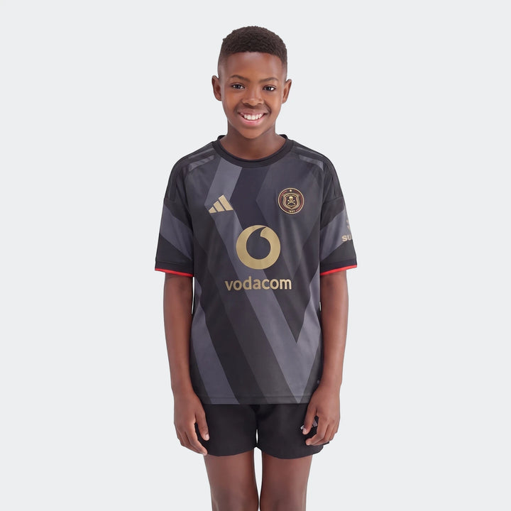 A young boy wearing the Orlando Pirates 26/27 Home Kids Jersey, featuring a black and grey pattern with gold logos.