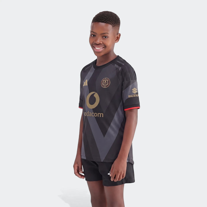Kids' Orlando Pirates 26/27 home jersey in black and grey chevron pattern with gold sponsor logos, worn by a smiling boy.