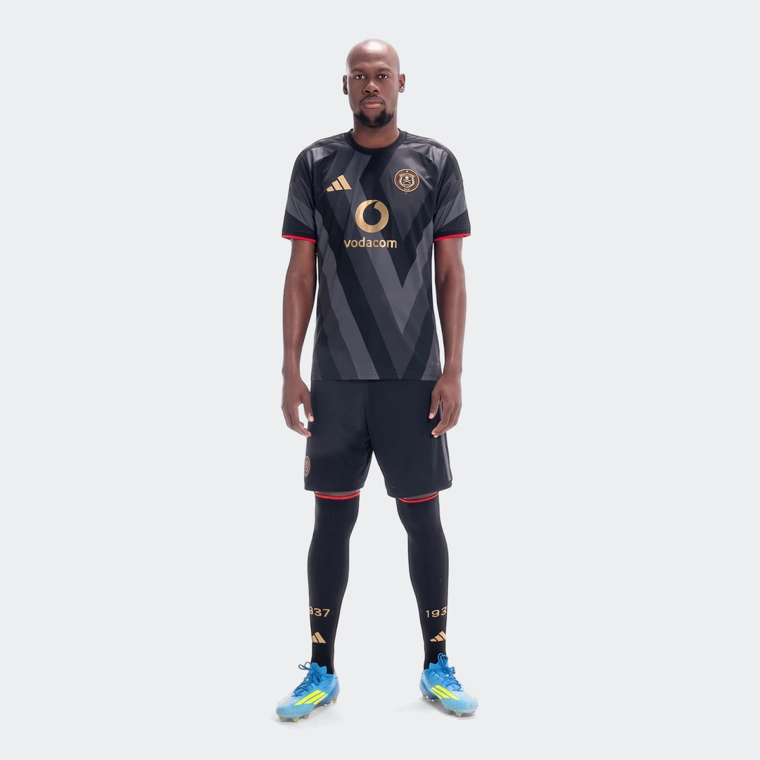 Male model wearing Orlando Pirates 26/27 Home Jersey in black and grey, with matching shorts, tights, and blue cleats.