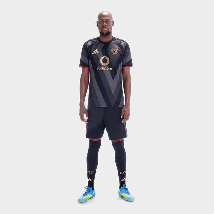 Male model wearing Orlando Pirates 26/27 Home Jersey in black and grey, with matching shorts, tights, and blue cleats.