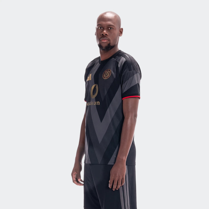 Male model wearing a black and gray Orlando Pirates 26/27 Home Jersey with gold logos, standing on a light background.