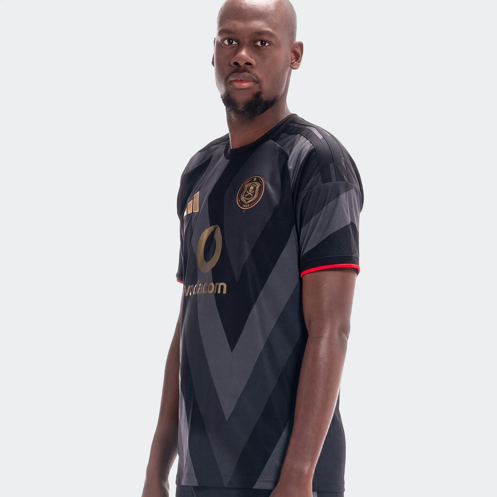Male model wearing an Orlando Pirates 26/27 Home Jersey, a black and grey soccer jersey with gold logos.