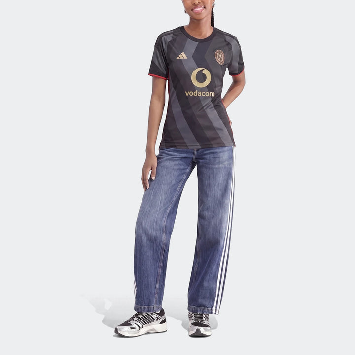 Young woman wearing Orlando Pirates 26/27 Home Jersey in dark grey with gold logos, styled with blue striped jeans and