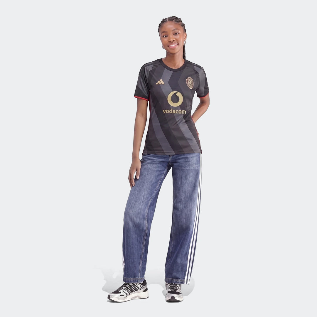 Young woman models the Orlando Pirates 26/27 Home Jersey, featuring a dark grey design with red trim and gold logos, paired