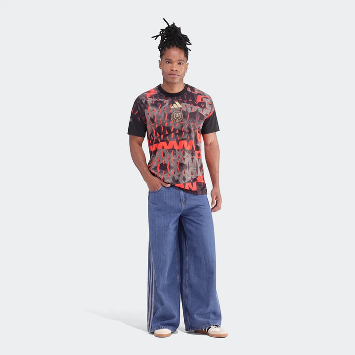 Male model in black and red patterned Orlando Pirates 26/27 Pre Match Jersey, wide-leg blue jeans, and sneakers.