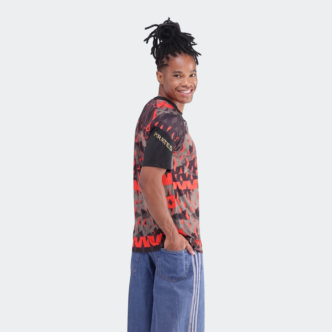 Smiling man wearing an Orlando Pirates 26/27 Pre Match Jersey in a red and black geometric pattern, paired with blue jeans,