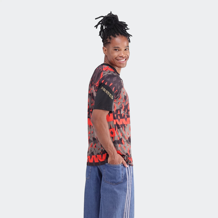 Smiling man wearing an Orlando Pirates 26/27 Pre Match Jersey in a red and black geometric pattern, paired with blue jeans,