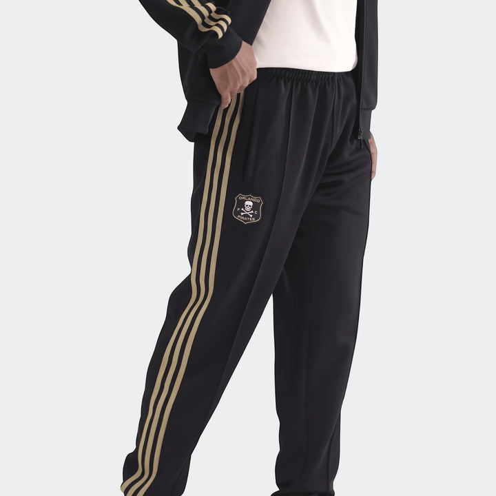 Orlando Pirates black track pants with gold stripes and club logo on the left thigh, worn by a person.