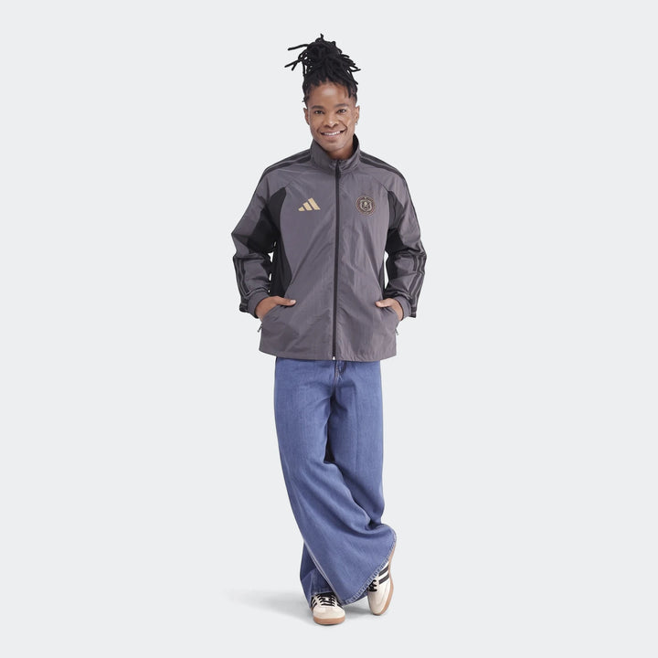 A full-body shot of a person wearing an Orlando Pirates Home Anthem Jacket in gray and black with gold logos and baggy blue