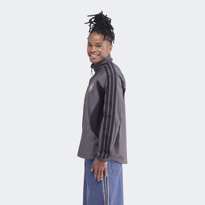 Man in a gray Orlando Pirates Home Anthem Jacket with black stripes and blue jeans, side profile view.