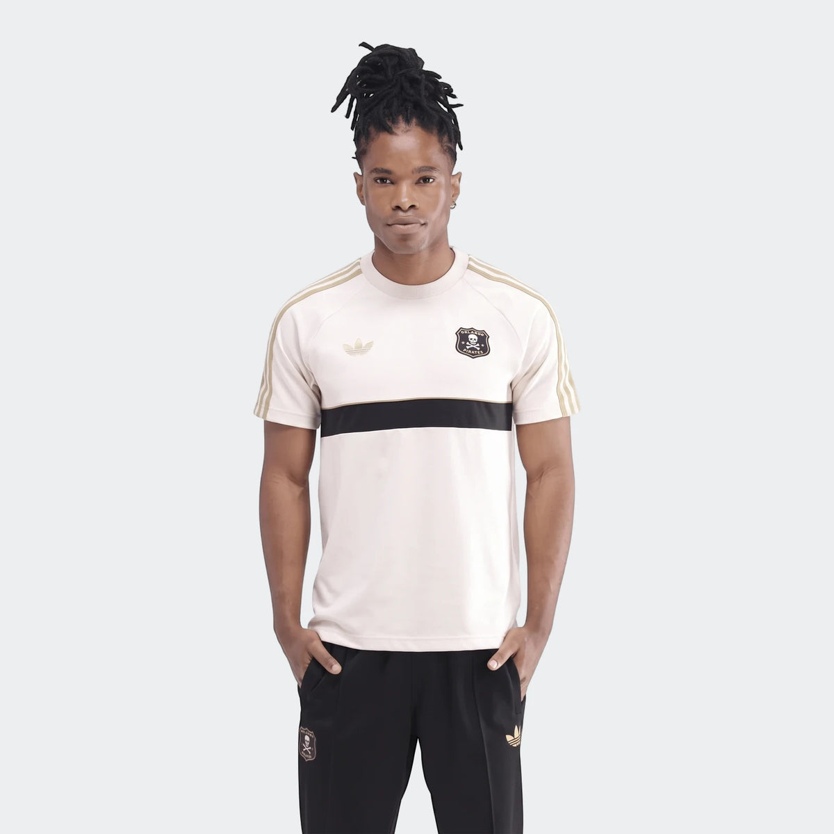 Male model wearing an Orlando Pirates Originals T-Shirt in cream with black and gold accents, paired with black Adidas track