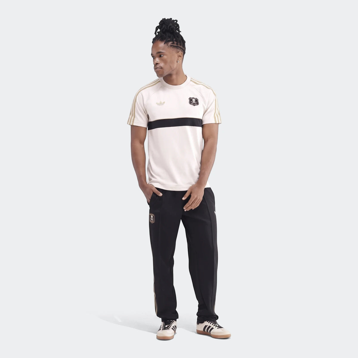 Male model in a white and black Orlando Pirates Originals T-Shirt with gold stripes and matching track pants, full body shot.