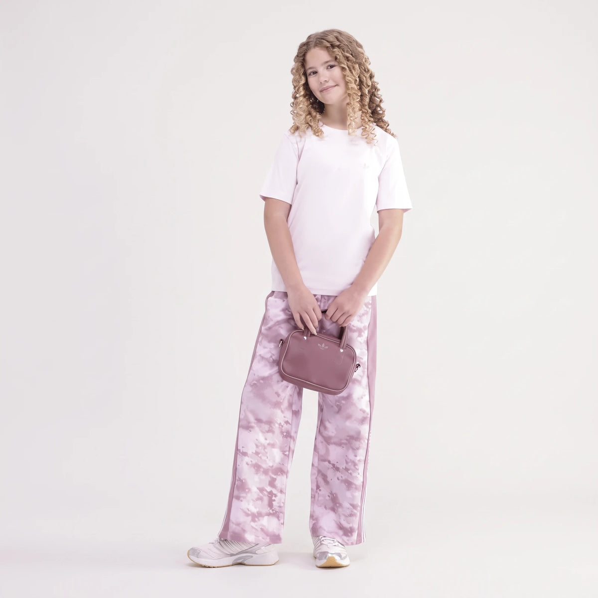 Young girl with curly hair wearing a pink Adidas Premium Essentials Tee, pink tie-dye track pants, and holding a small pink