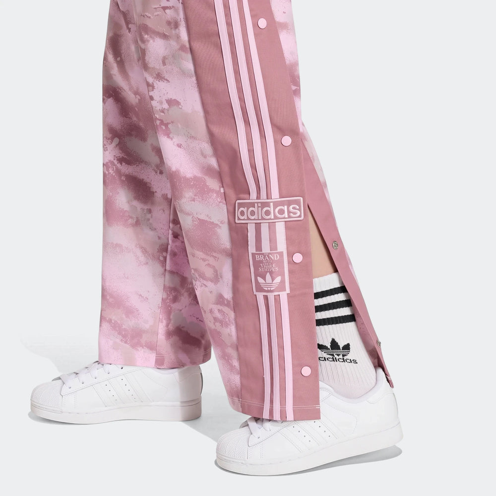Close-up of a person wearing pink tie-dye Adidas Loose Adibreak Camo Twill Trousers and white sneakers, showing side snap