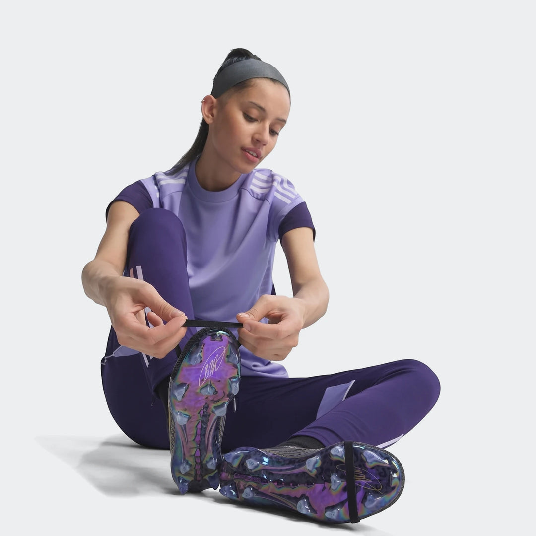 A woman sits, tying iridescent Predator Elite Ft Fg Kaka Edition football boots, wearing a purple athletic outfit.