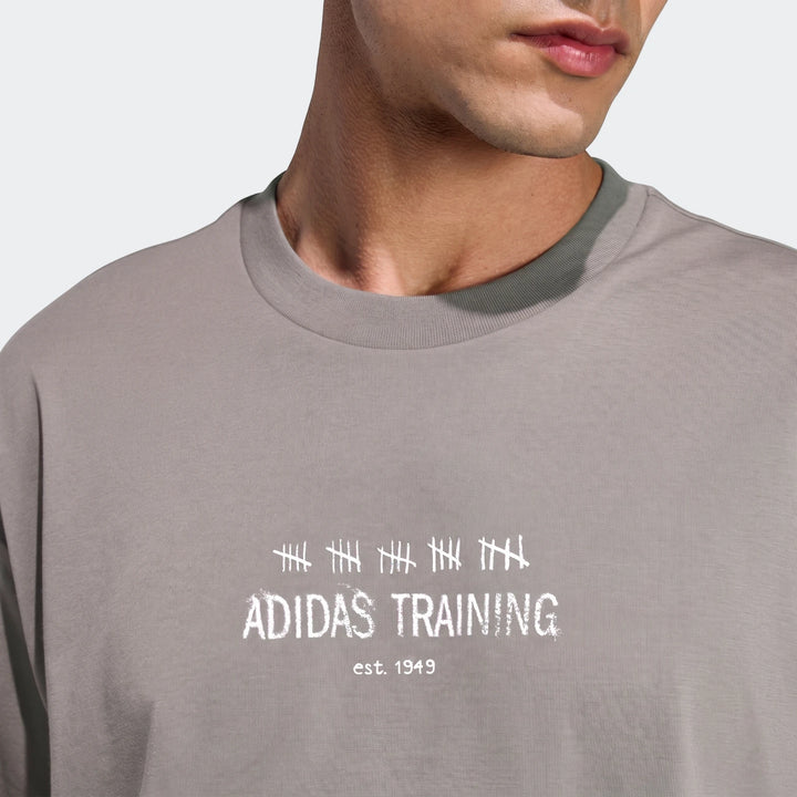 Male model wearing a grey Adidas Primelift Graphic T-shirt with white logo print, shown in a close-up studio shot.