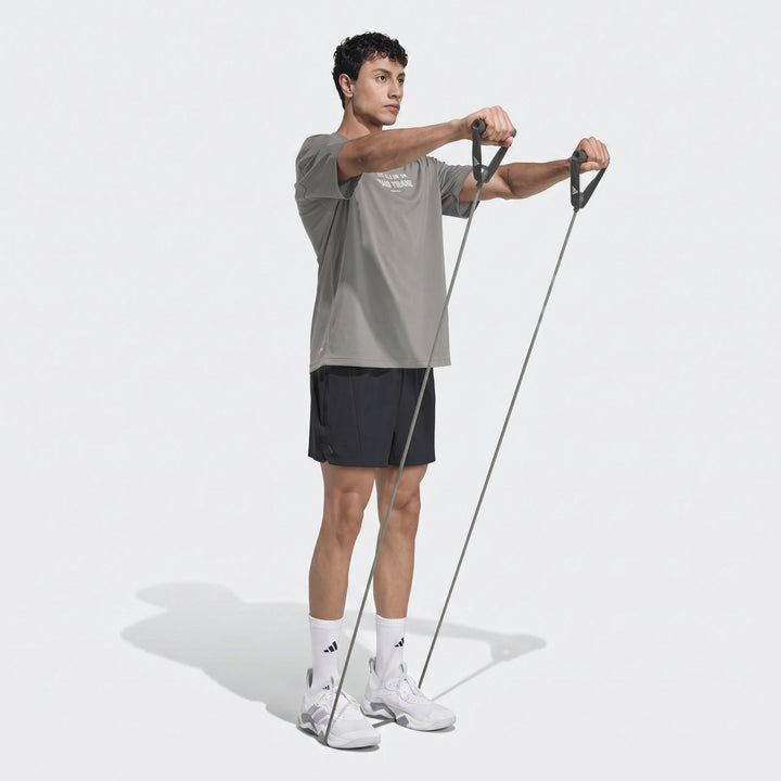 A man wears a grey Primelift Graphic T-Shirt and black shorts, using a resistance band in a full-body studio shot.
