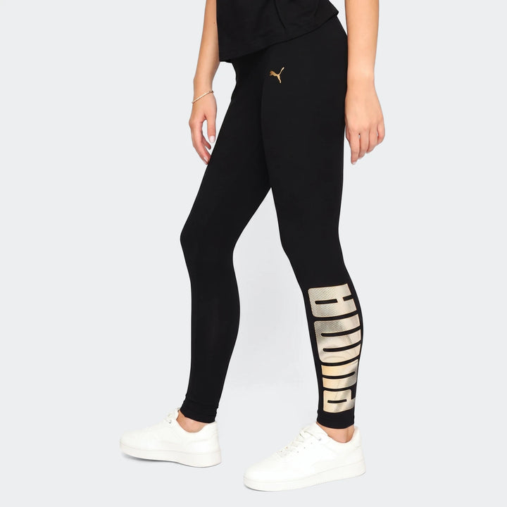 Model wearing black Puma Ess Metallic Leggings with gold logo detailing and white sneakers, side view.