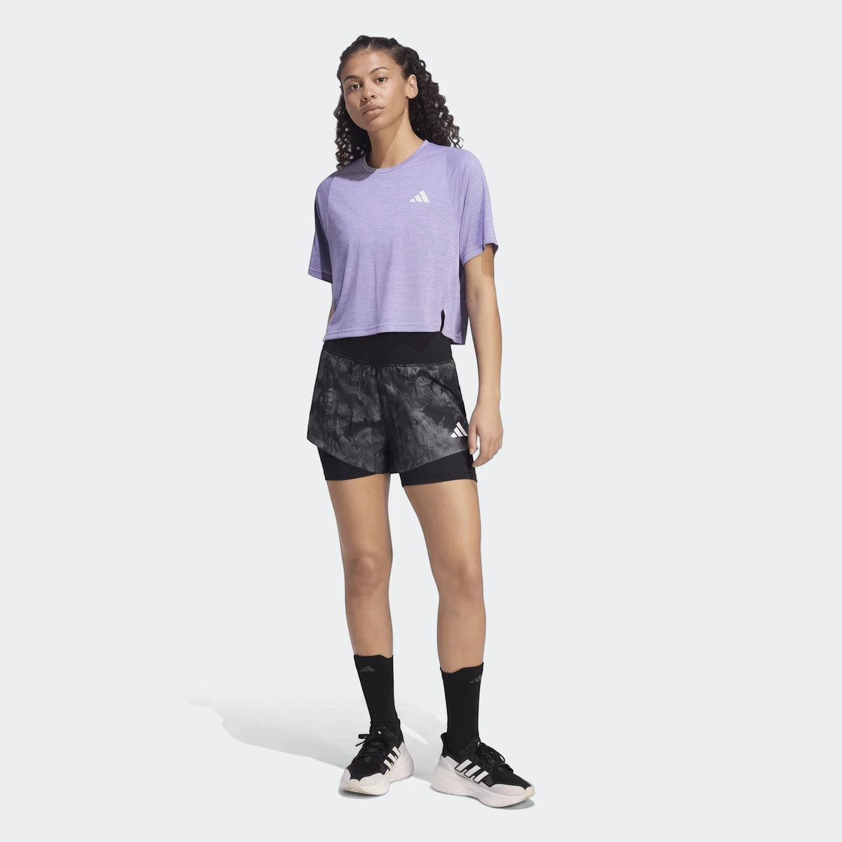 Woman wearing a purple Adidas Adi365 Soft Touch T-Shirt and black patterned shorts, standing full body.