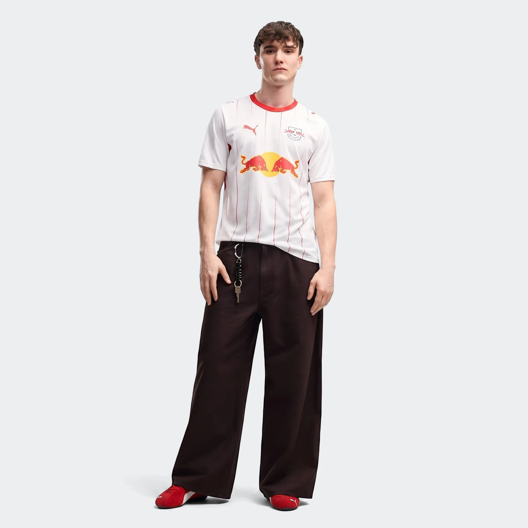 Men's Rb Leipzig 26/27 Home Jersey in white with red stripes, worn by a male model with dark pants and red shoes.