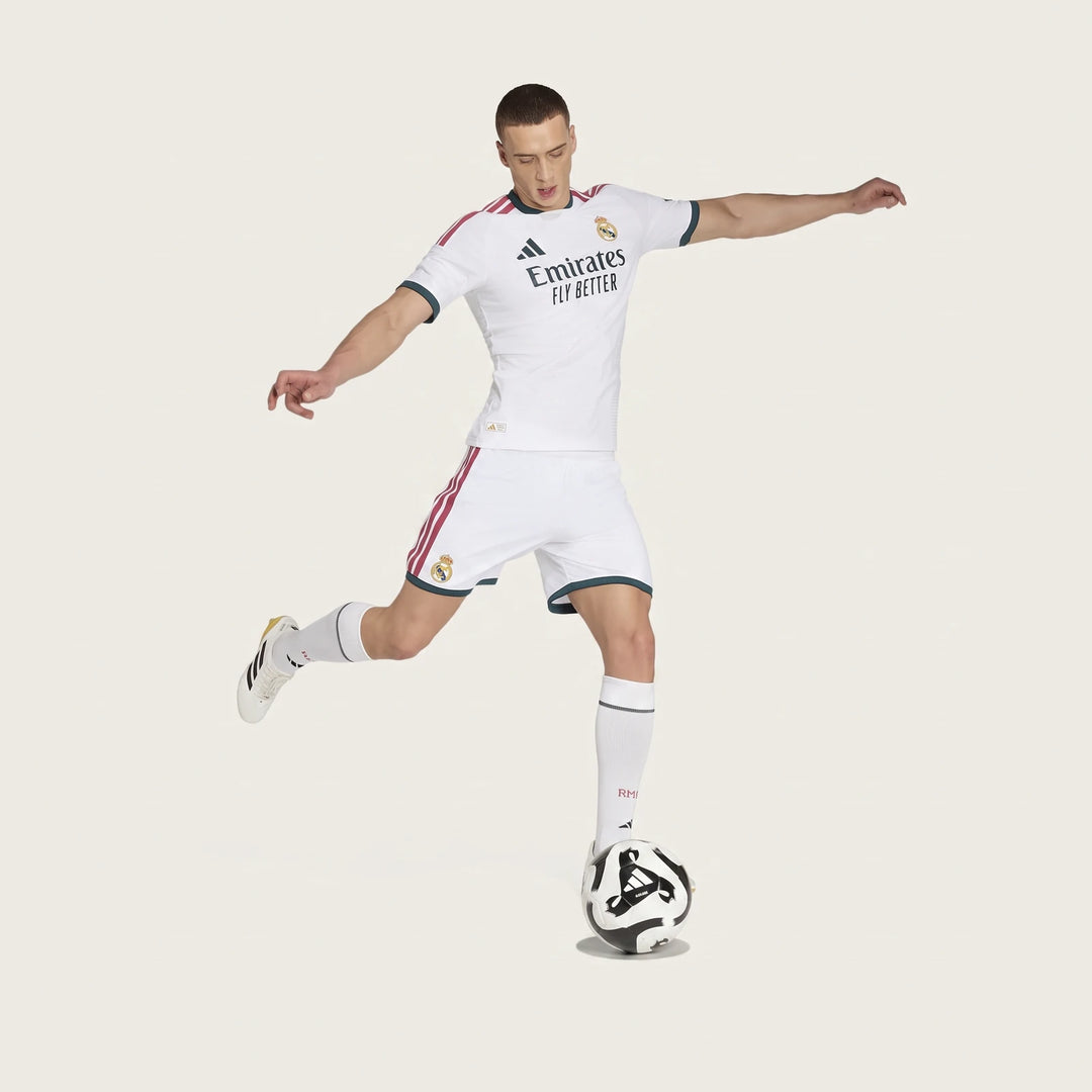 Male model in a white Real Madrid 25/26 Authentic Home Jersey and matching shorts, featuring red stripes and green trim,