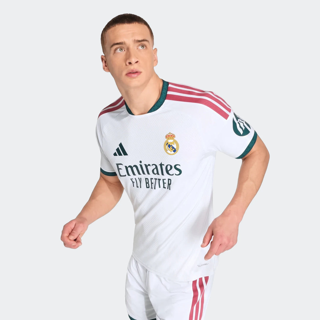 Male model wearing a white Real Madrid 25/26 Authentic Home Jersey with red shoulder stripes and green trim.