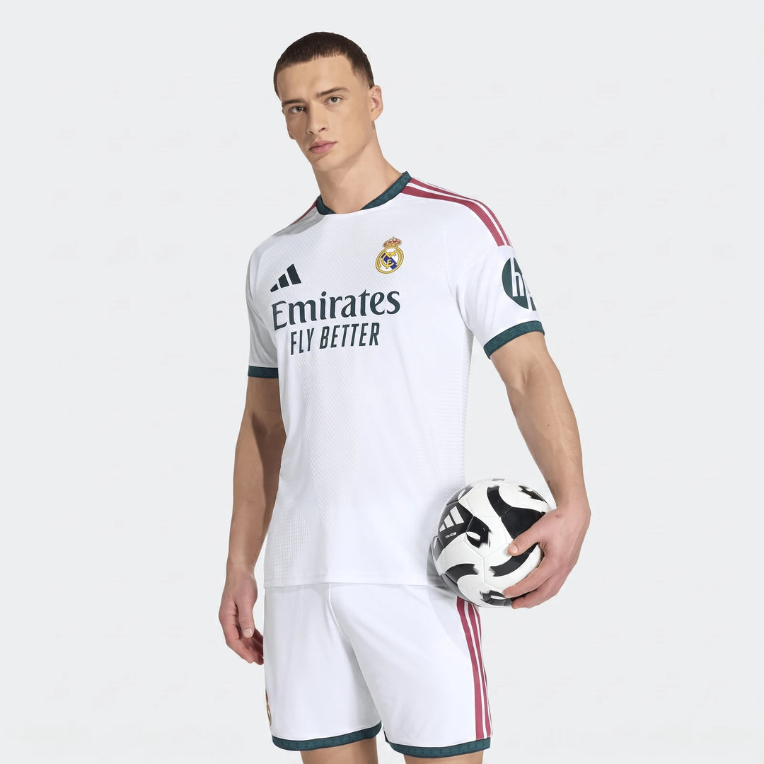 Male model wearing a white Real Madrid 25/26 Authentic Home Jersey and white shorts, holding a black and white soccer ball.