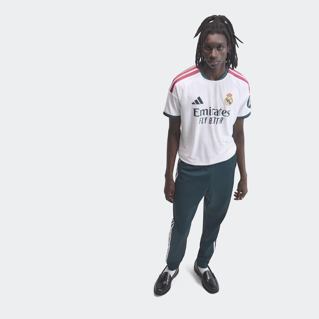 Male model wearing a white Real Madrid 25/26 Authentic Home Jersey with red shoulder stripes and dark green track pants,