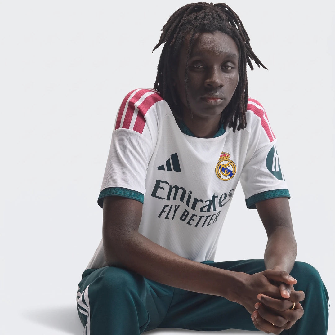 Real Madrid 25/26 Authentic Home Jersey in white with red stripes and green trim, worn by a male model sitting.