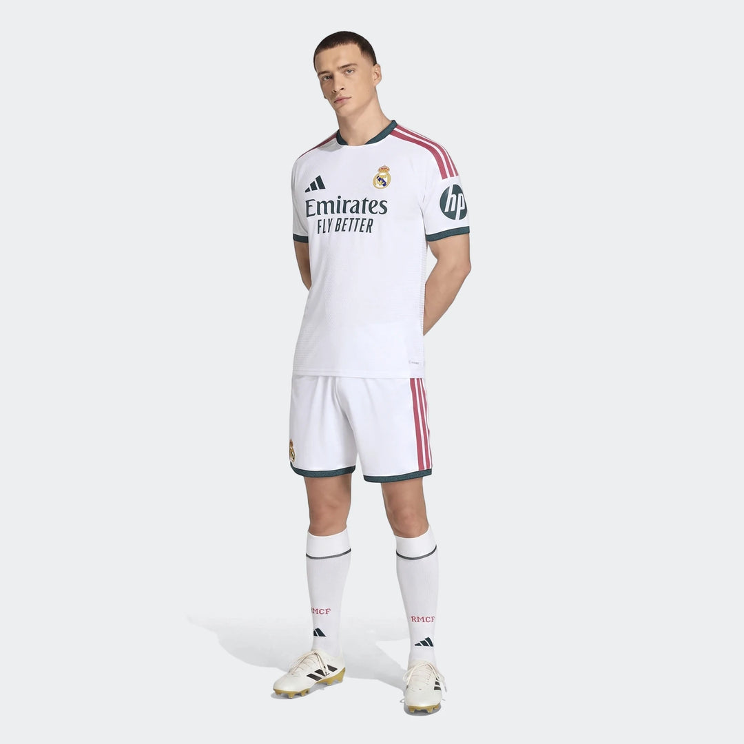 Male model wearing a white Real Madrid 25/26 Authentic Home Jersey with red stripes and dark green trim, white shorts, and