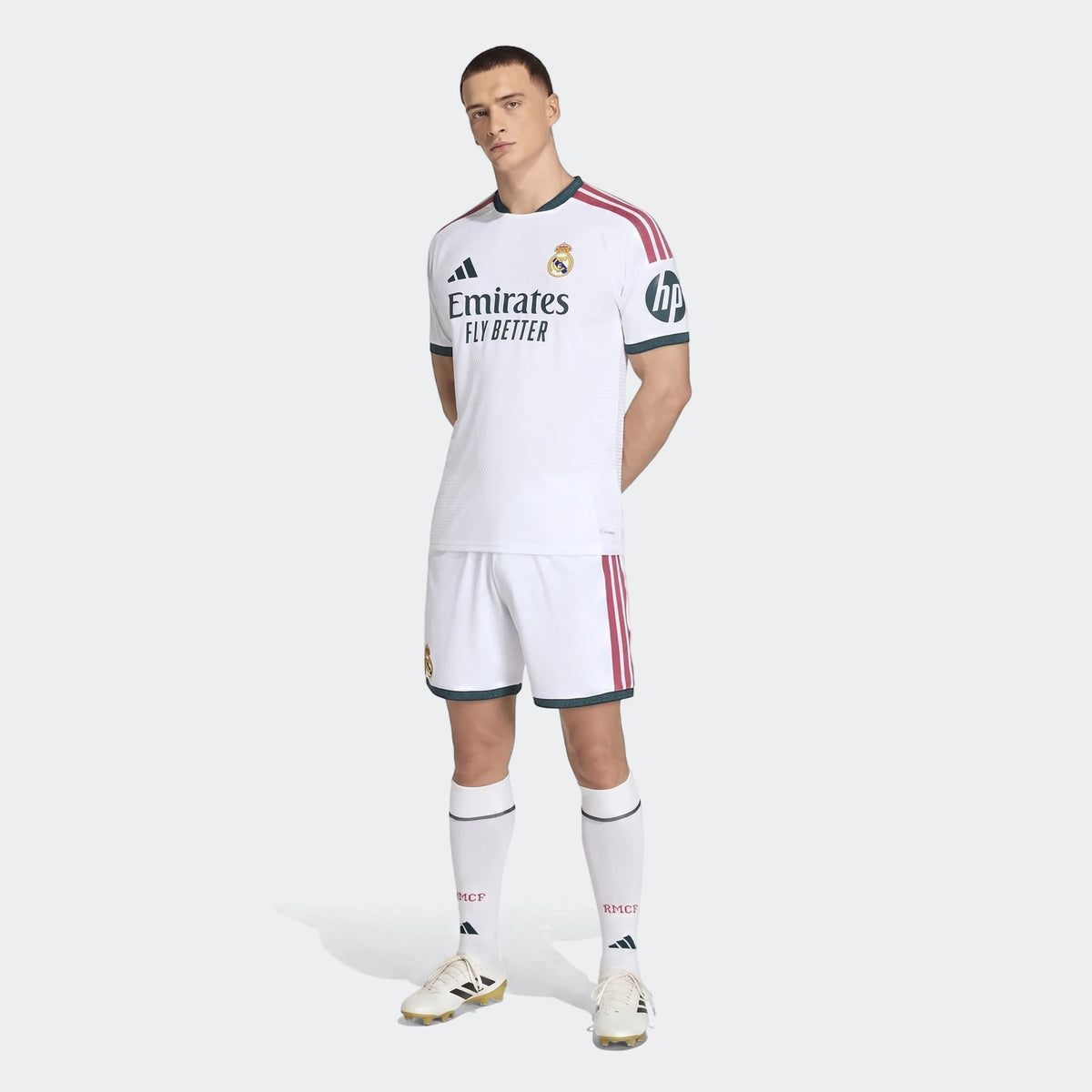 Male model wearing a white Real Madrid 25/26 Authentic Home Jersey with red stripes and dark green trim, white shorts, and