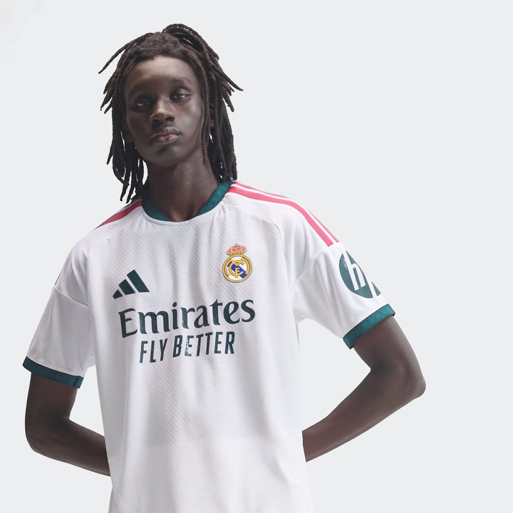Male model wearing a white Real Madrid 25/26 Authentic Home Jersey with green and pink trim and sponsor logos.