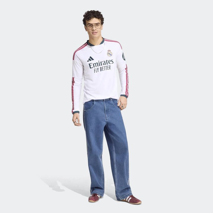 A male model wears a white Real Madrid 25/26 long-sleeve home jersey, blue wide-leg jeans, and red sneakers.